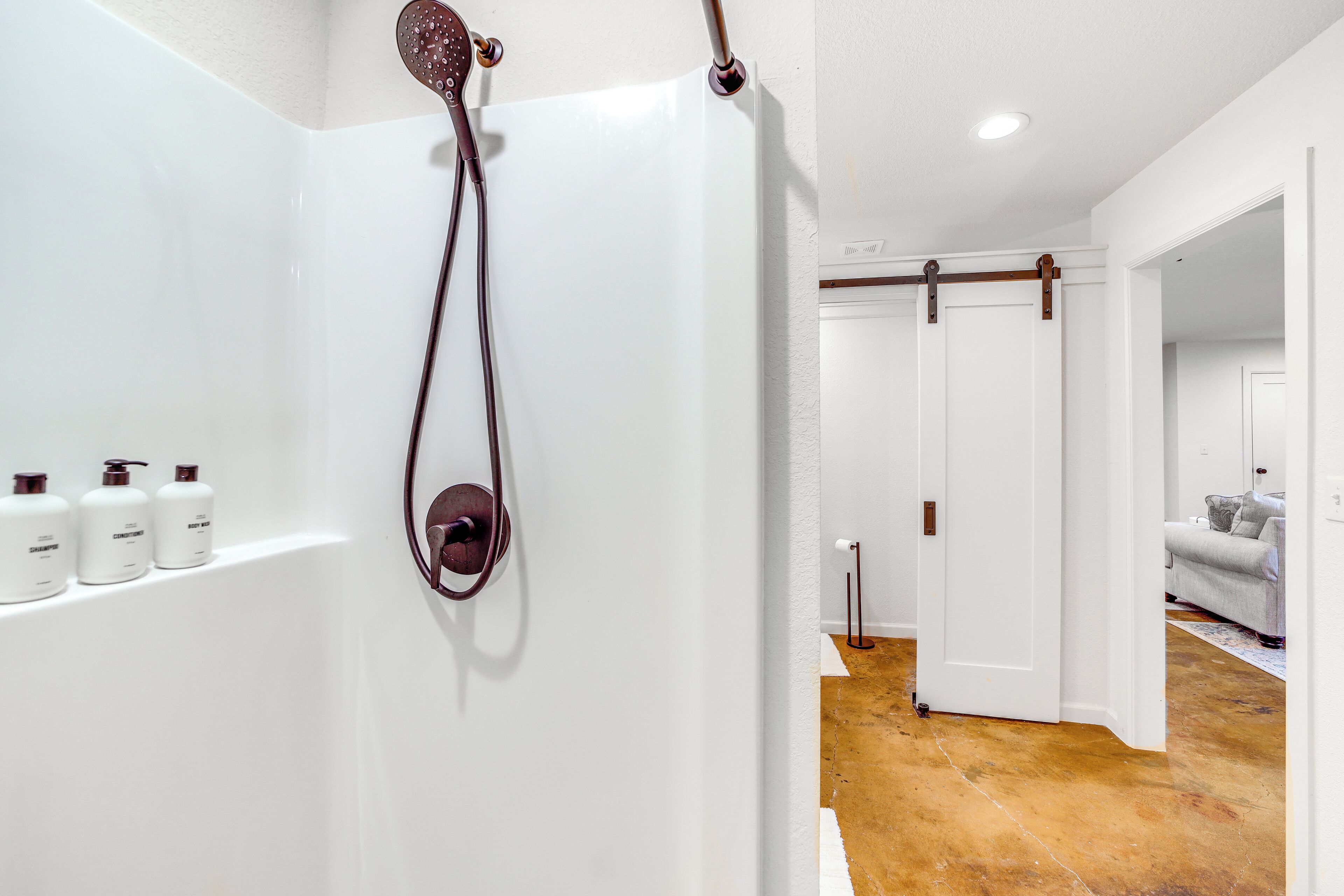 A bathroom with a shower, white walls, and a wooden floor. There are three bottles of shampoo and conditioner on the wall.