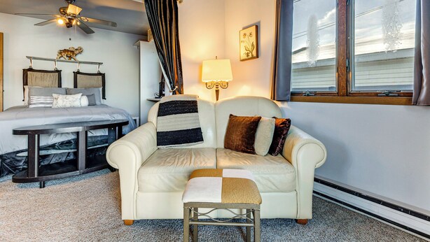 Une chambre confortable avec un lit, un canapé, une petite table, un ventilateur de plafond, une fenêtre avec des rideaux et une lampe fixée au mur.