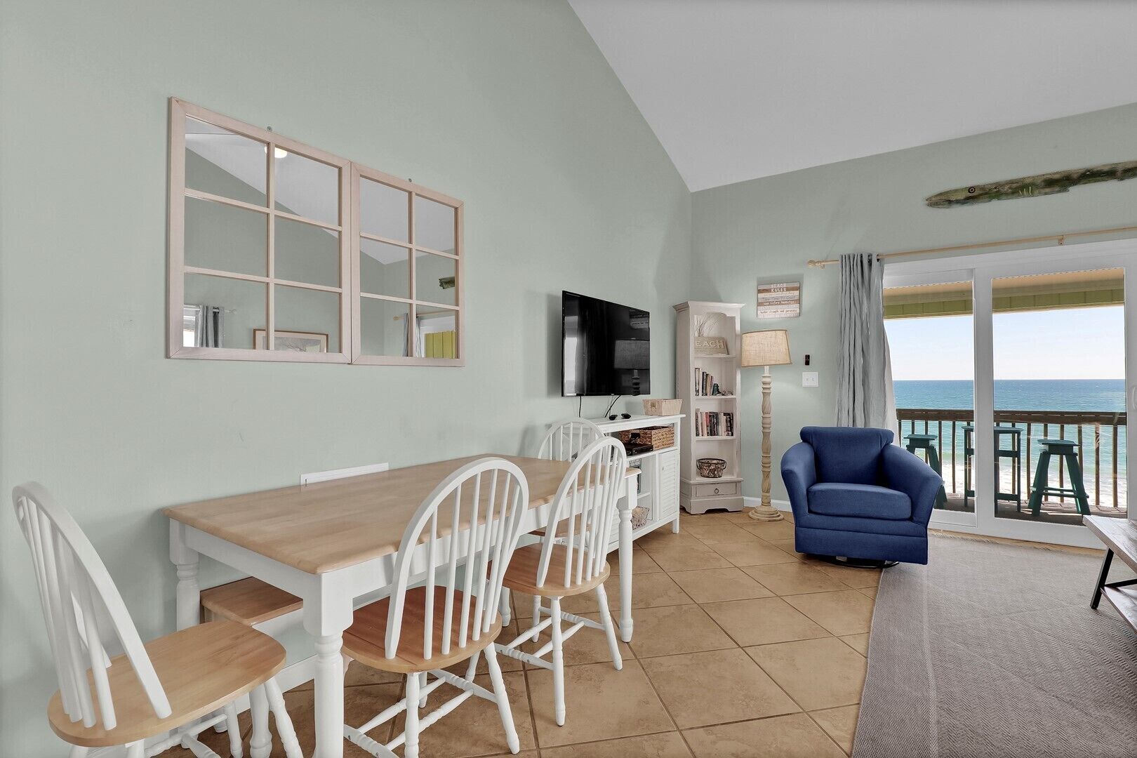 A dining area with a wooden table and white chairs, a television, a bookshelf, and a view of the ocean through a balcony.