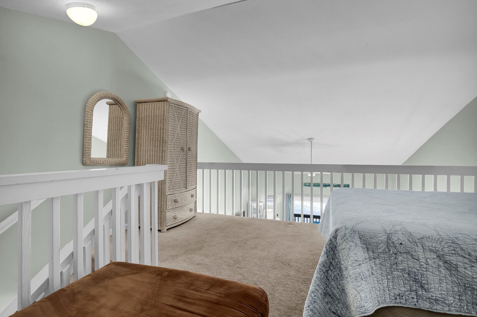 A bedroom with a sloped ceiling, a white railing, a brown bench, a wicker wardrobe, and a bed with a patterned blanket.