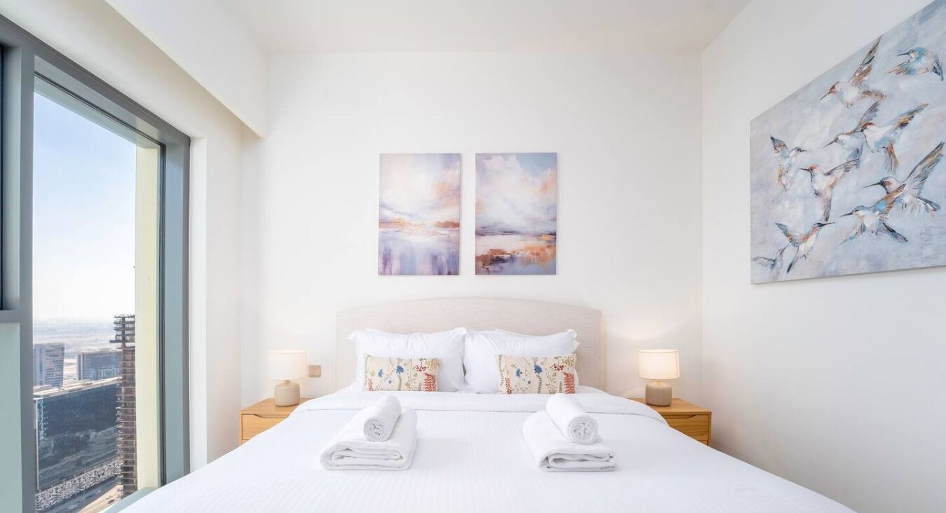 Et hotelværelse med en stor seng, to billeder hængt på væggen, et vindue med udsigt over byen og et natbord med en lampe.