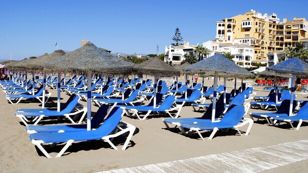 Une plage bordée de rangées de chaises longues bleues et d’ombrelles en chaume.