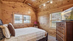 A cozy wooden cabin bedroom with a bed, dresser, and two windows offering views of the outdoors. - Spectacular Home w Hot Tub Fireplace Jacuzzi (Sevierville)