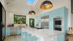 A modern kitchen with light blue cabinets, stainless steel appliances, a large island with bar stools, and pendant lighting. - Kamalani Modern in Princeville (Princeville)