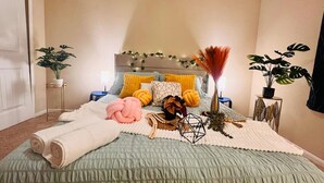 A neatly arranged bedroom with a bed, pillows, a bedside table, a lamp, and two potted plants. - Spacious 4BR Home w Game Room Office Backyard (Kyle)