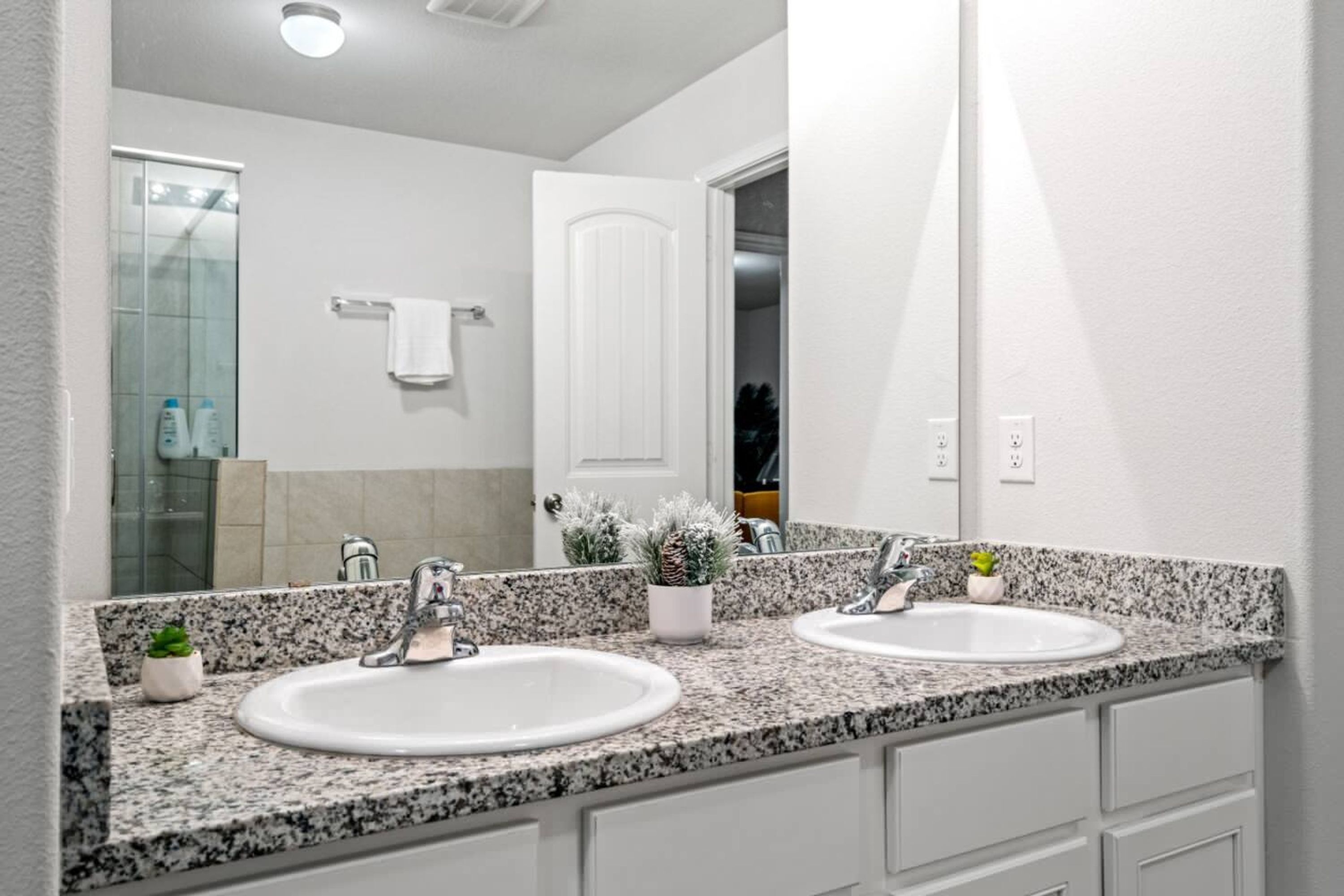 A bathroom with two sinks, granite countertops, a large mirror, and a door leading to another room.
