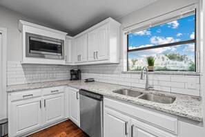 A modern kitchen with white cabinets, a stainless steel dishwasher, a double sink, and a window with a view of trees.