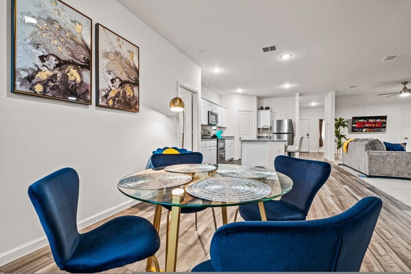 A modern living room with a glass dining table surrounded by blue chairs, a kitchen in the background, and artwork on the walls. - One Level Home Foosball Firepit Quiet Neighborhood (Lavon)