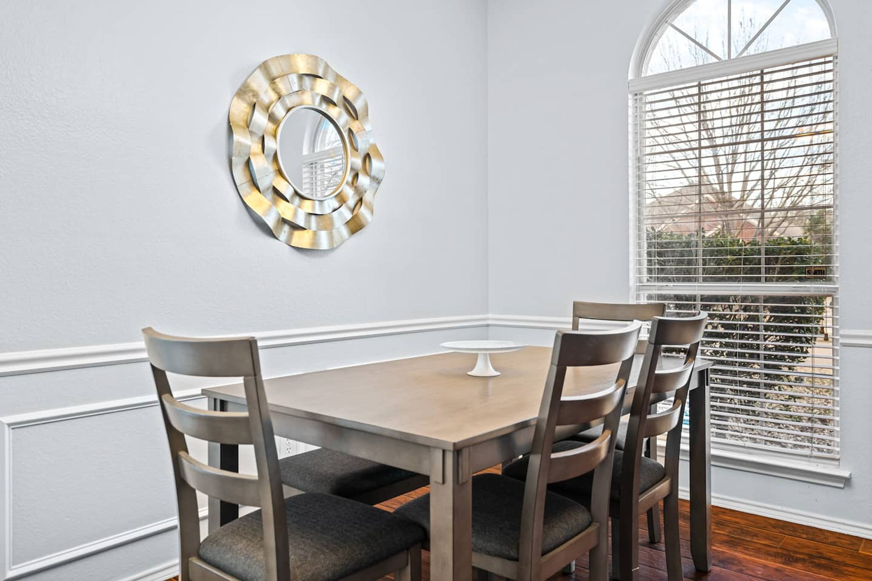 A dining room with a wooden table and four chairs, a decorative mirror, and a window with blinds.