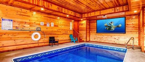 An indoor swimming pool with wooden walls, a pool rule sign, a lifebuoy, and a television screen displaying a frog.