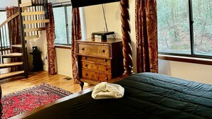 A bedroom with a bed, a dresser, a television, a spiral staircase, and a window with a view of trees.