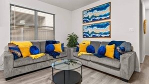 Una sala de estar con dos sofás grises, cojines amarillos y azules, una mesa de centro de vidrio y dos cuadros abstractos en la pared.