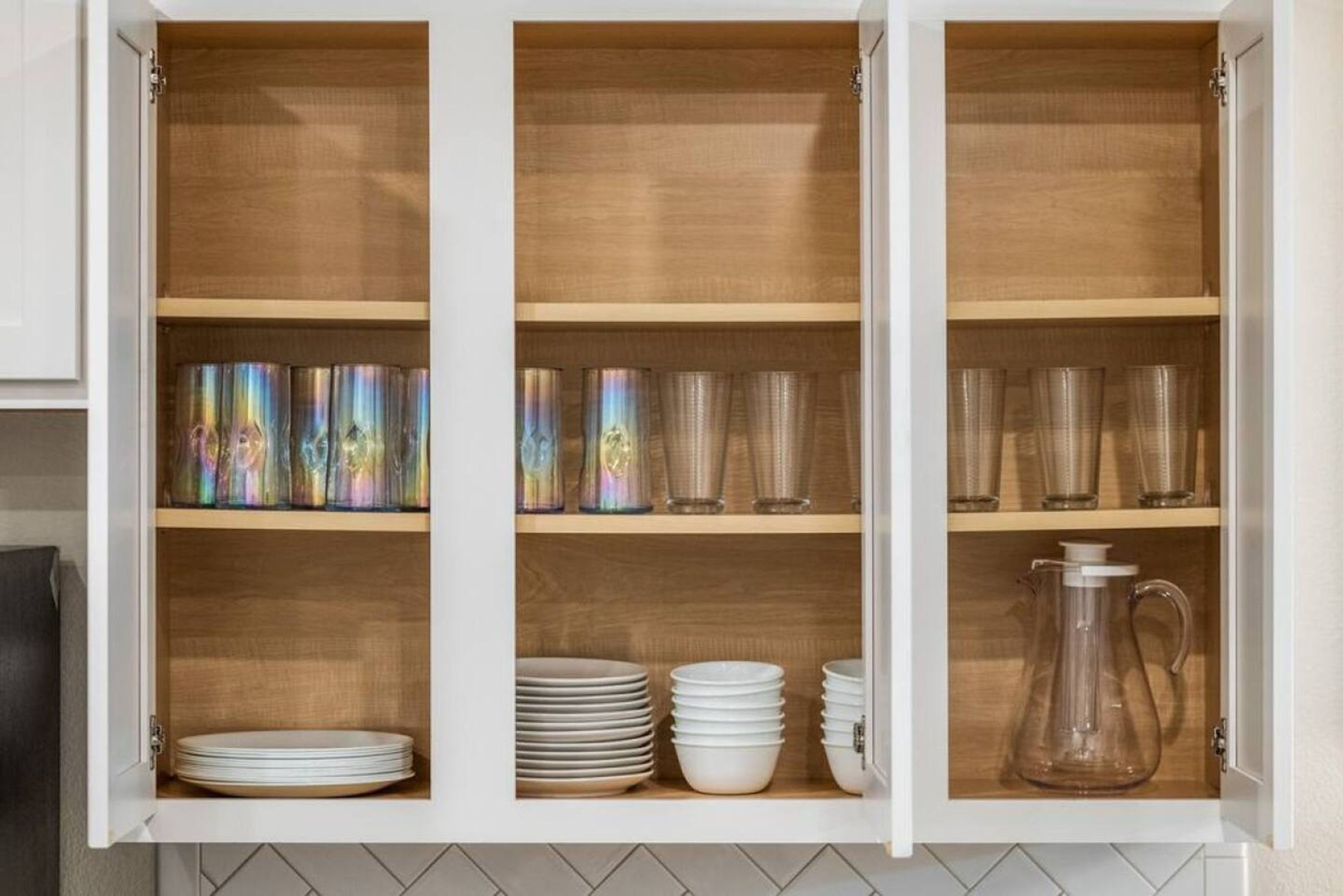 Ein Küchenschrank mit drei Einlegeböden, auf denen Teller, Gläser und eine Karaffe stehen.