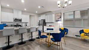 Cocina moderna con armarios blancos, microondas de acero inoxidable y estufa a gas. Hay taburetes de bar y una mesa redonda con sillas azules.