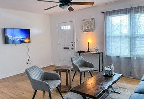 A living room with a television, two armchairs, a coffee table, a ceiling fan, a door, and a window with curtains. - Affordable 2BR Getaway Near Plaza District Lilac1 (Midwest City)