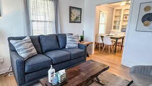 A living room with a grey sofa, a wooden coffee table, and a dining area with a wooden table and chairs. - Affordable 2BR Getaway Near Plaza District Lilac1 (Midwest City)