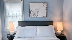 A bedroom with a bed, two bedside lamps, a window with blinds, and a wall-mounted abstract painting. - Affordable 2BR Getaway Near Plaza District Lilac1 (Midwest City)