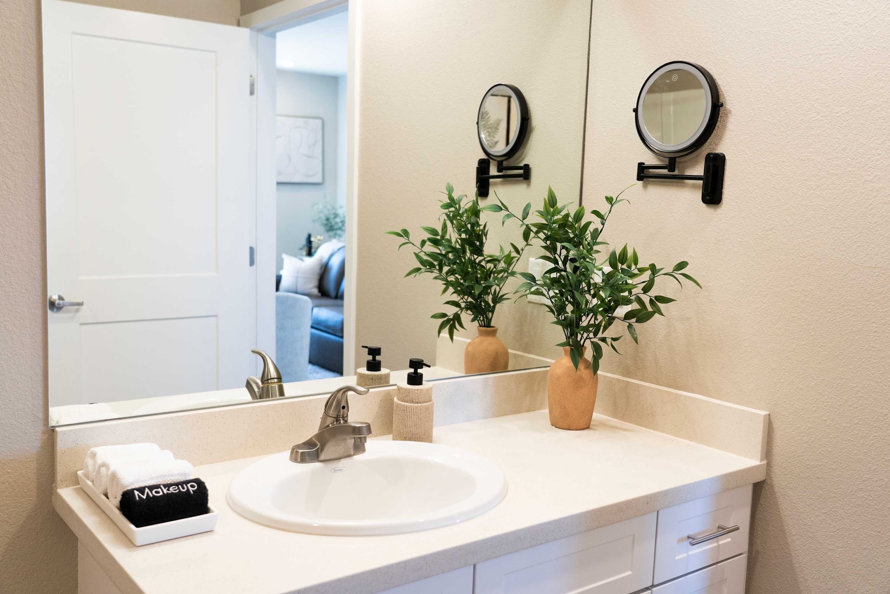 Une salle de bain avec un lavabo blanc, deux miroirs, un porte-serviettes sur lequel est posée une serviette étiquetée « Maquillage », et deux plantes en pot.
