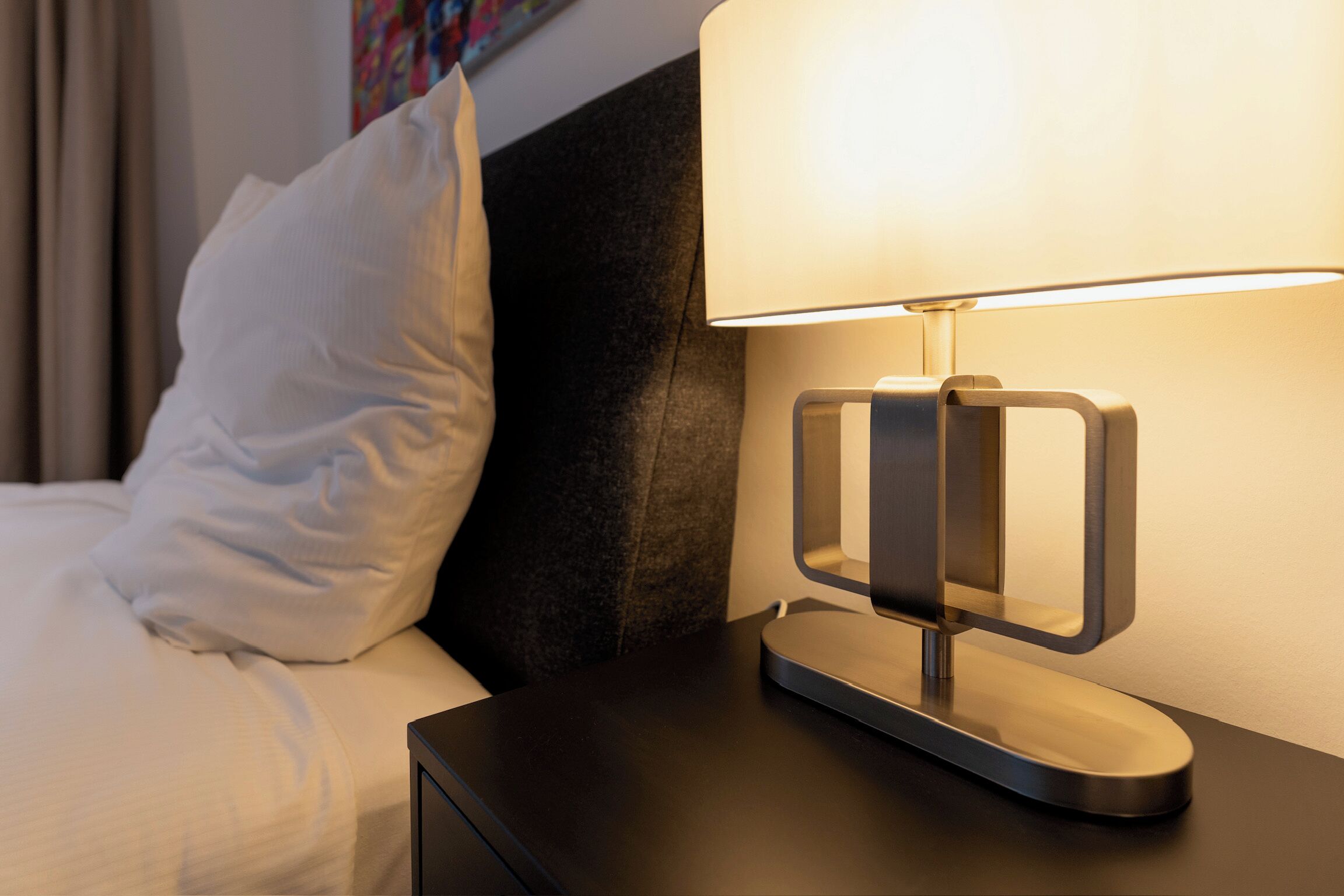 Un lit bien fait, avec une literie blanche, une tête de lit sombre et une lampe de chevet moderne posée sur une table de chevet noire.