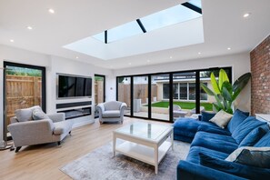 Una sala de estar moderna con techo de cristal, un sofá azul, un sillón gris y un televisor.