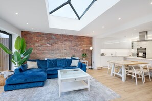 A modern living room with a blue sectional sofa, a white coffee table, and a dining area with a wooden table and white chairs. - Sustainable Stays (Bushey)