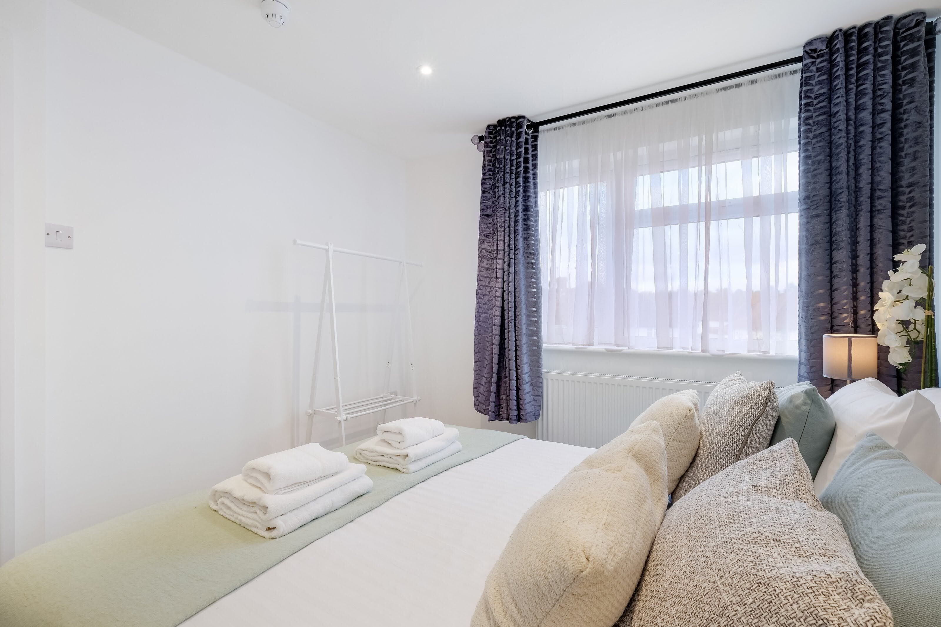Ein Hotelzimmer mit einem Bett, einem Heizkörper, einem Fenster mit Vorhängen sowie einem Nachttisch mit Lampe und Blumen.