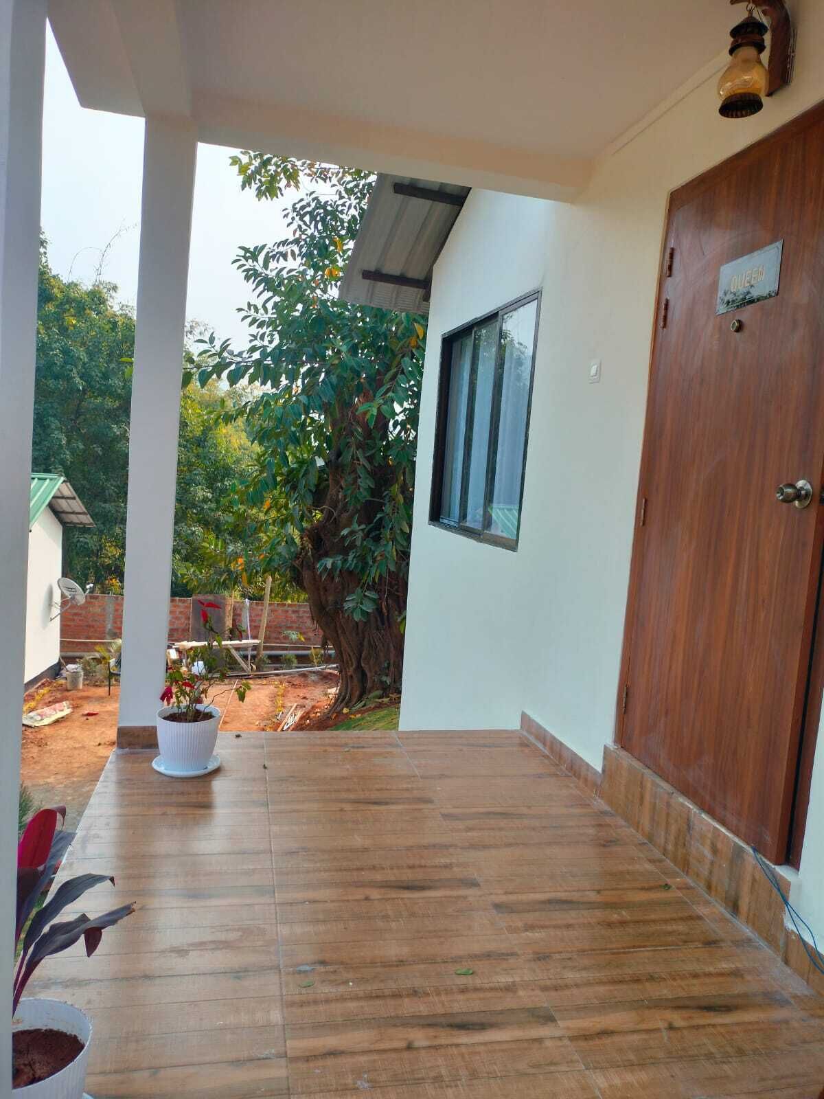 A covered porch with a wooden floor, a potted plant, and a view of a garden with trees and a brick wall.