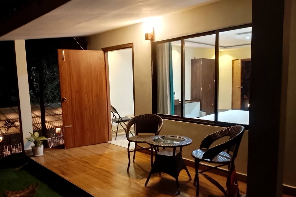 Un área de estar al aire libre con sillas de mimbre y una mesa redonda pequeña, una puerta de madera, y vista del interior a través de un ventanal amplio.
