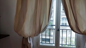 Una camera da letto con un letto, una finestra con tende, un termosifone e vista sugli edifici.