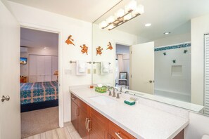Une salle de bain avec un grand miroir, un lavabo et une douche. Des serviettes sont accrochées au mur, et une petite étagère contient des articles de toilette.