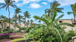 A tropical garden with palm trees, a wooden bridge, and a building with a beige facade. - Maui Kamaole K-207 Recently Remodeled, Views, Pools, Hot Tub, A/C, W/D (Kihei)