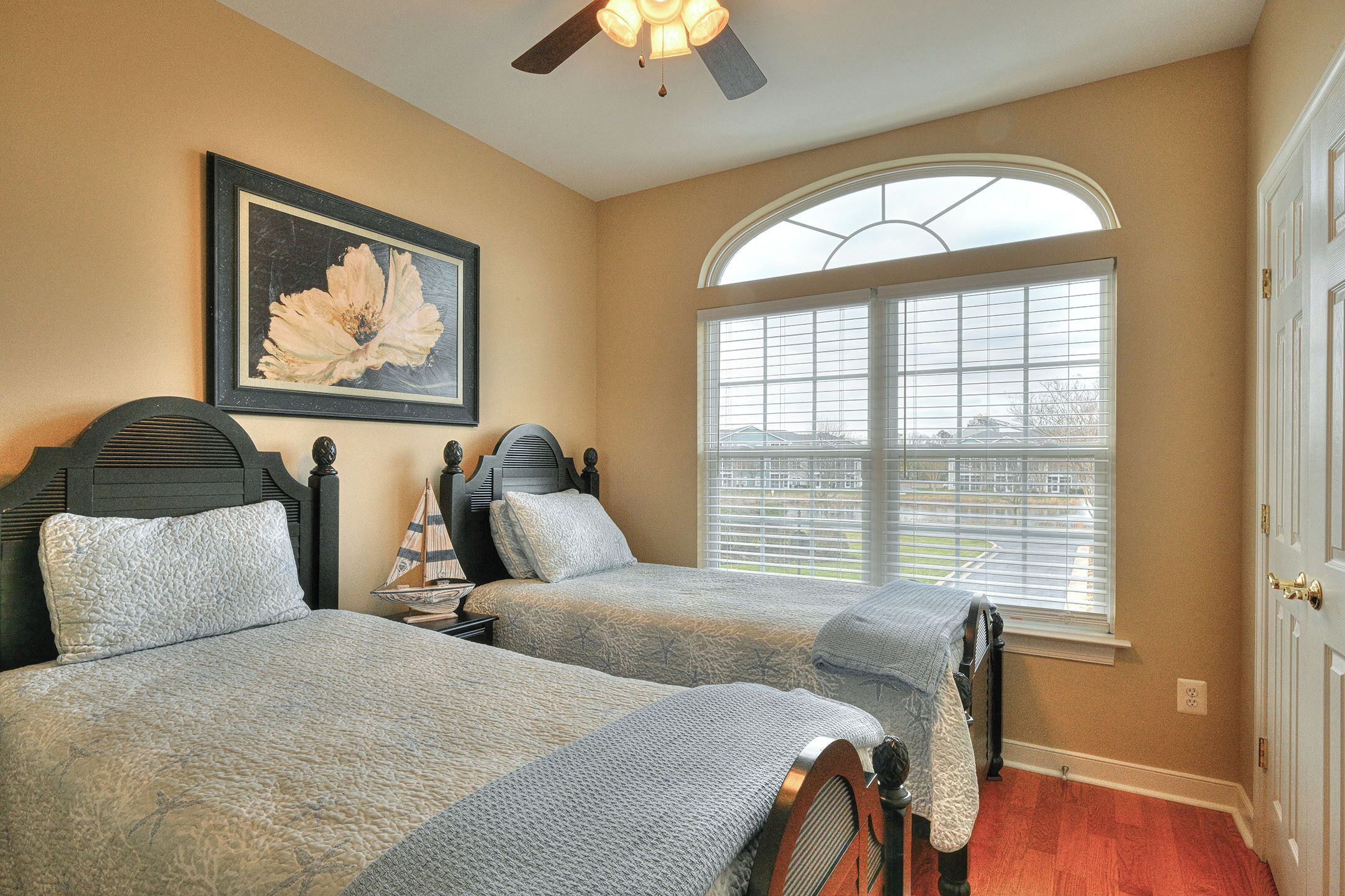A bedroom with two beds, a large window with blinds, a ceiling fan, and a framed picture on the wall.