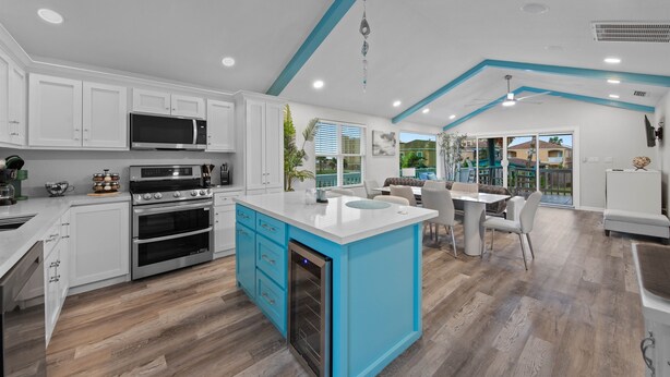 A modern kitchen with white cabinets, a blue island, stainless steel appliances, and a dining area with a view of the outdoors.