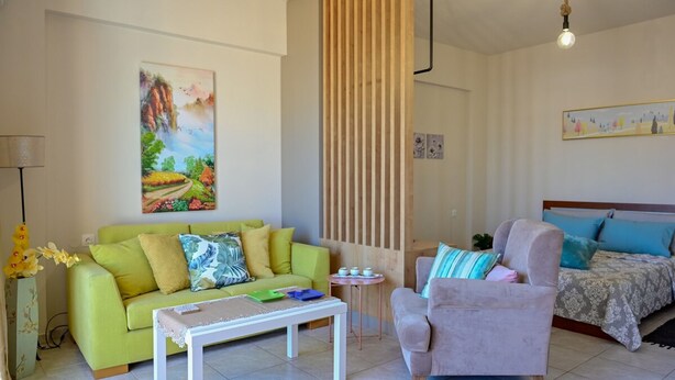 Ein Wohnzimmer mit einem grünen Sofa, einem grauen Sessel, einem weißen Couchtisch, einem Bett mit blauer Bettwäsche und einer Holzwand.