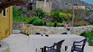 An outdoor dining area with a table and chairs, surrounded by stone walls and greenery.