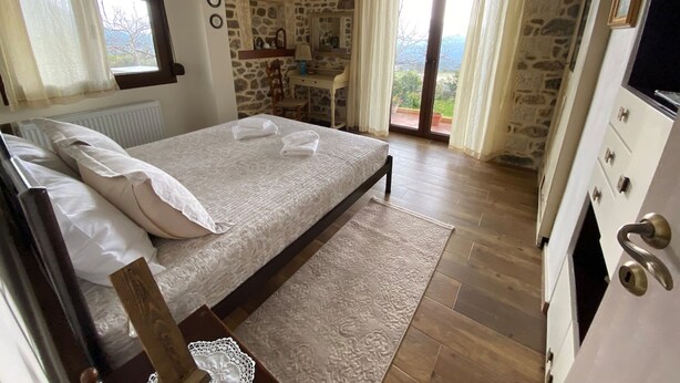 A bedroom with a stone wall, a bed with white linens, a wooden nightstand, a dresser, and a view of the outdoors.