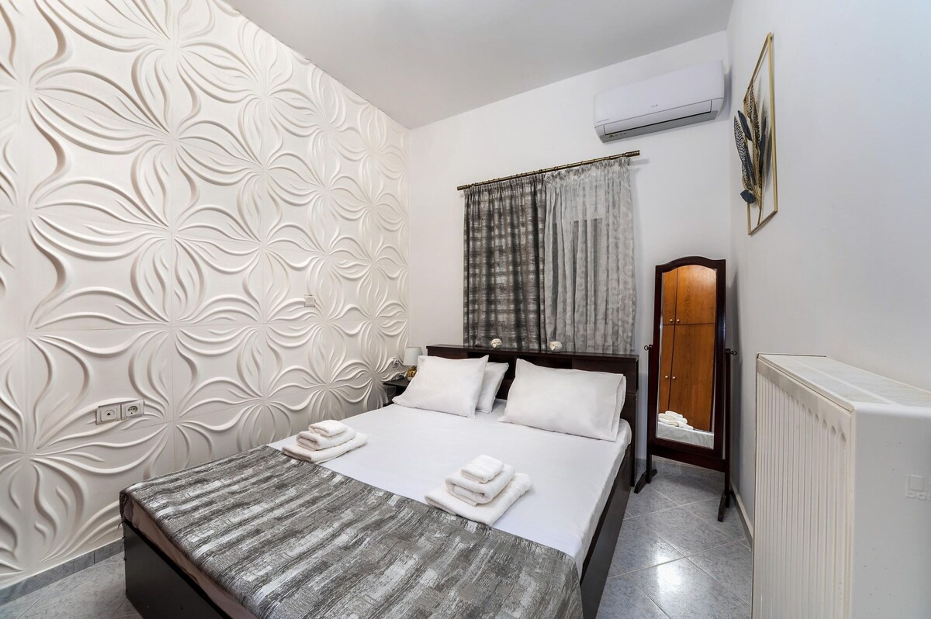Una cama bien hecha con sábanas y toallas blancas, cabecera de madera, un radiador y una pared con estampado floral.