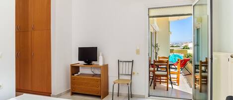 A bedroom with a bed, a TV on a wooden cabinet, a chair, and a balcony with a table and chairs.