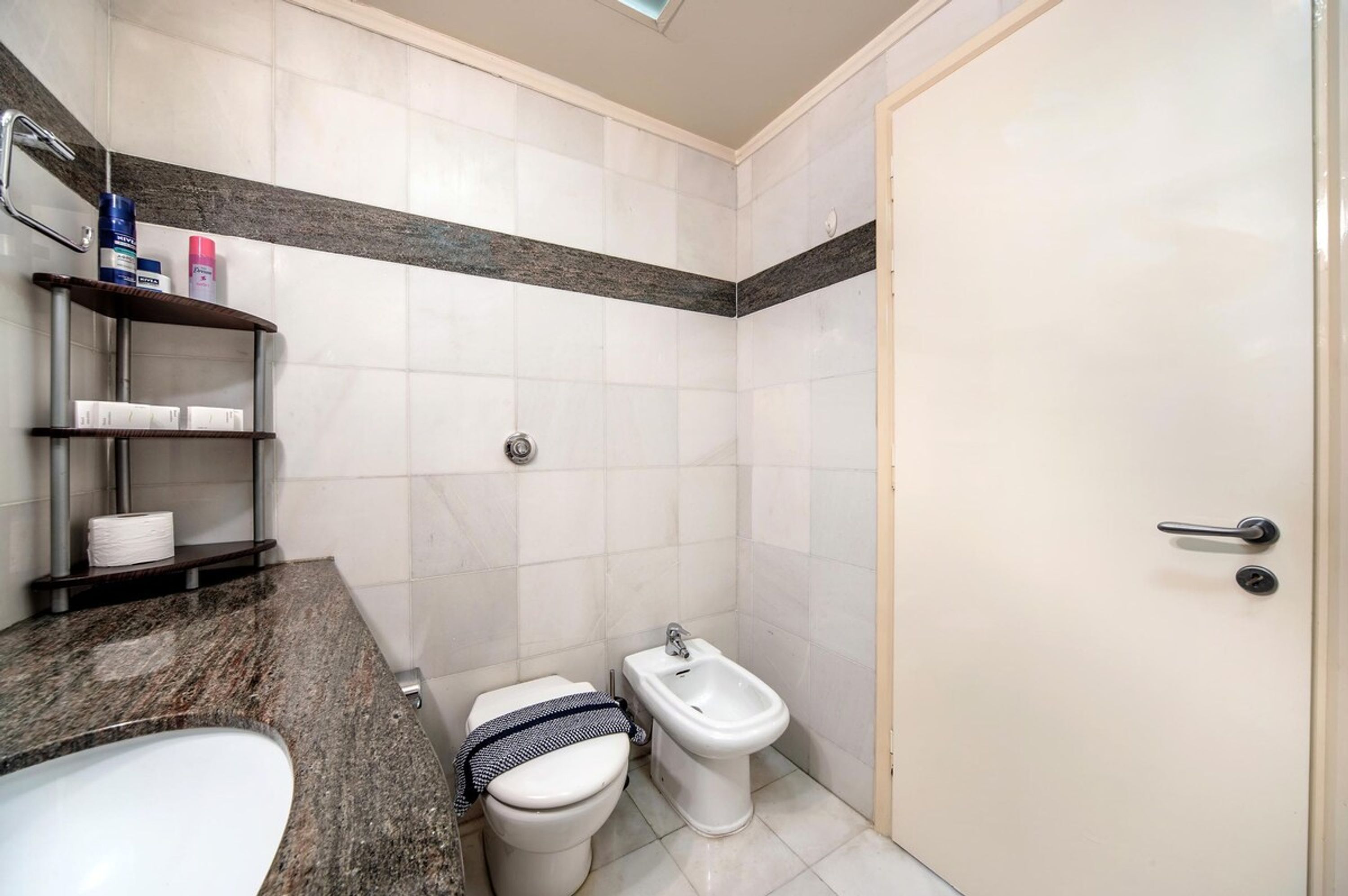 Un baño con inodoro, bidé y lavamanos. Hay estantes con artículos de aseo y un perchero para toallas.