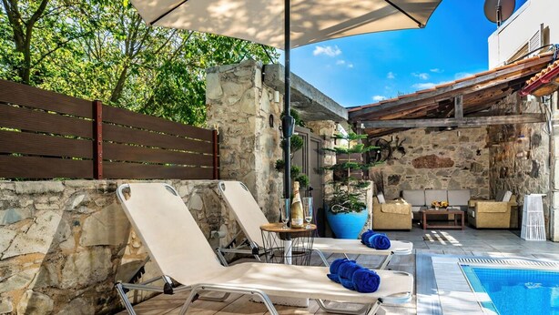A poolside area with lounge chairs, a table with a bottle and glass, and a stone wall. - Villa Kalouas Private Pool (Malevizi)