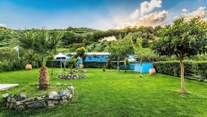 A lush green garden with a stone wall, a palm tree, and a blue building with a white roof. - Villa Kalouas Private Pool (Malevizi)