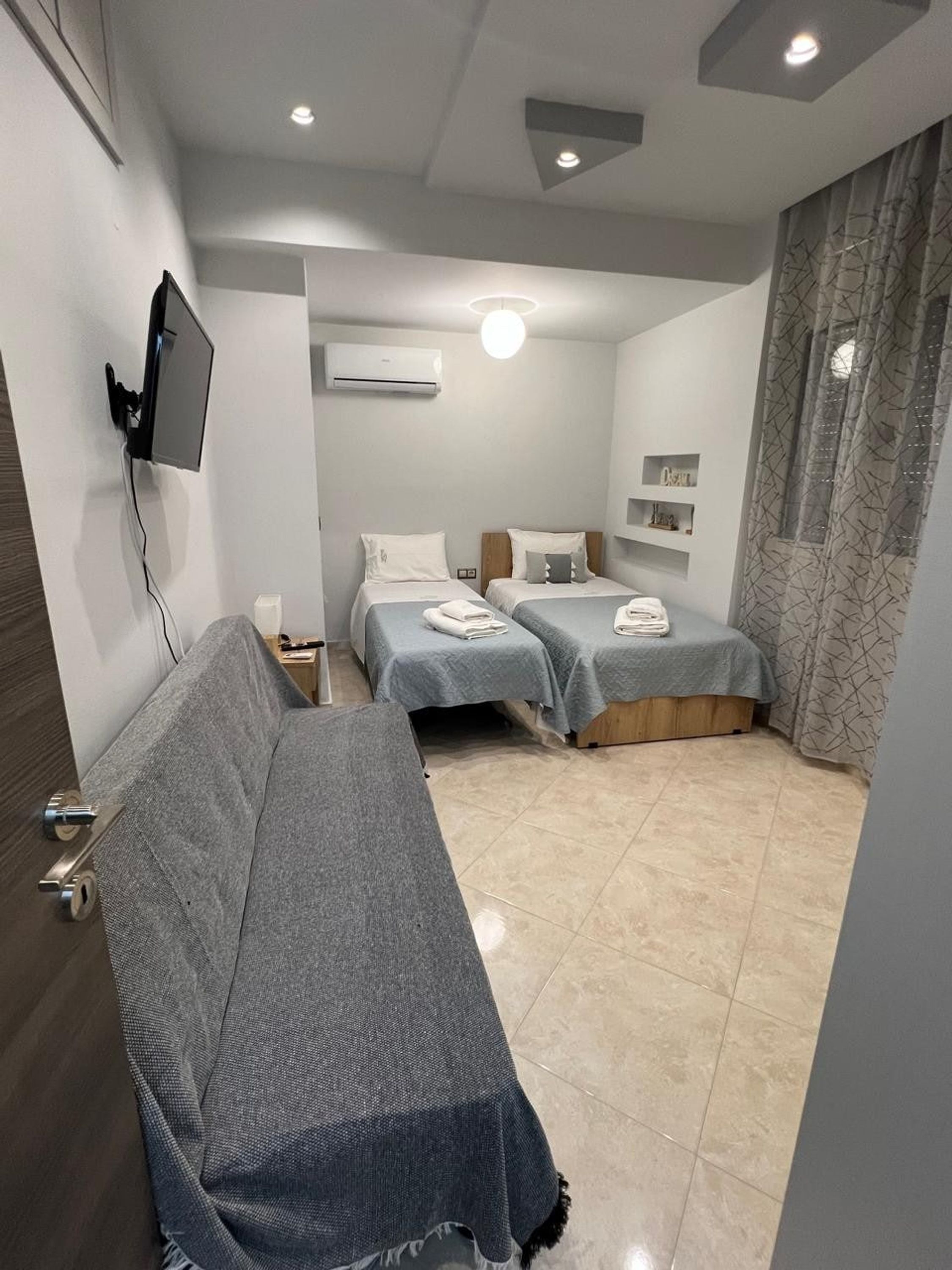A modern hotel room with two single beds, a sofa, a flat-screen TV mounted on the wall, and a ceiling light.