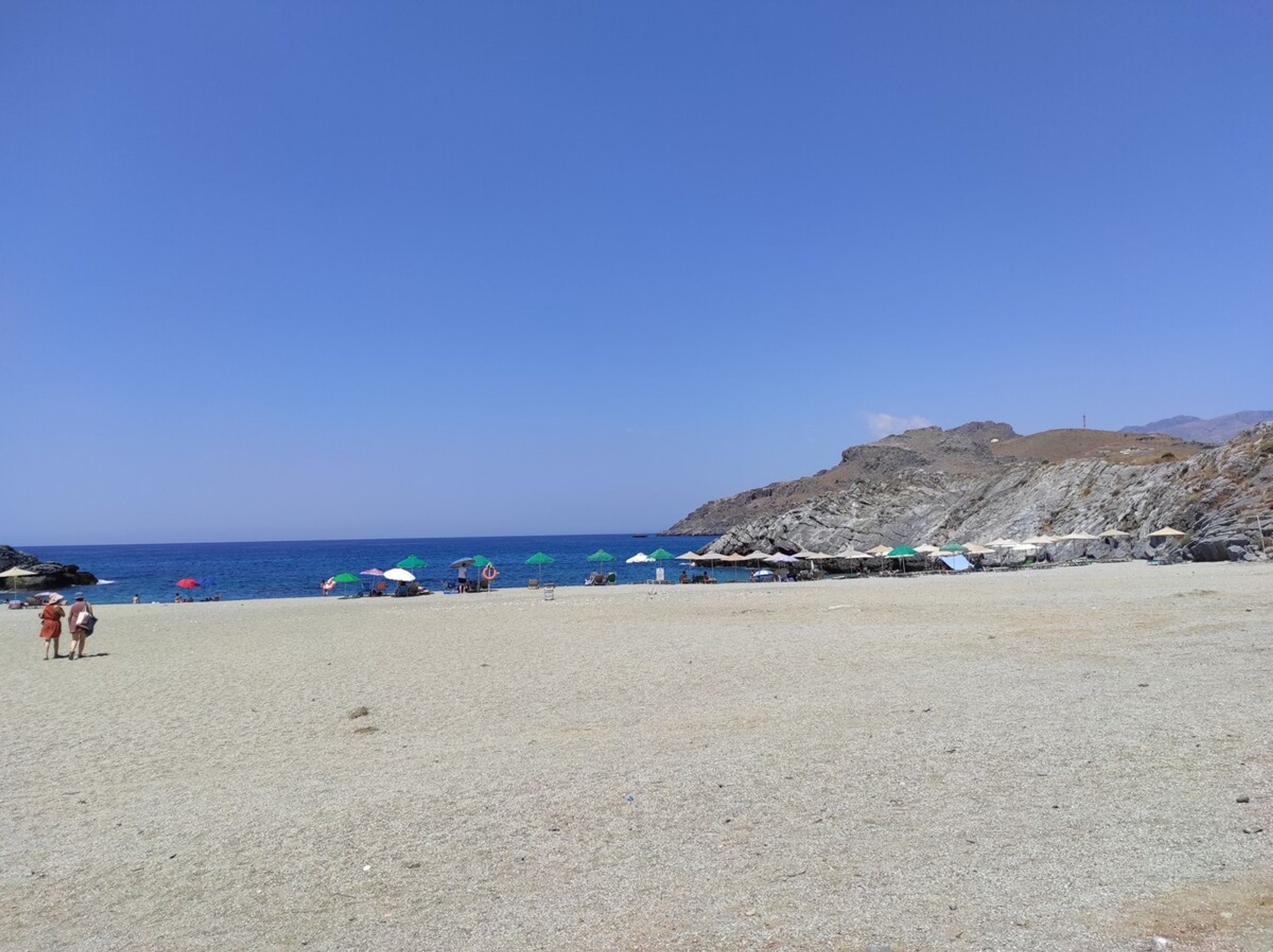 Una playa con un afloramiento rocoso al fondo, cielos despejados y varios sombrillas.