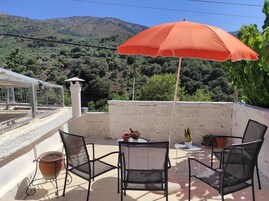 Un balcón con dos sillas, una mesa y un paraguas naranja. Hay plantas en macetas y se ve un paisaje montañoso.