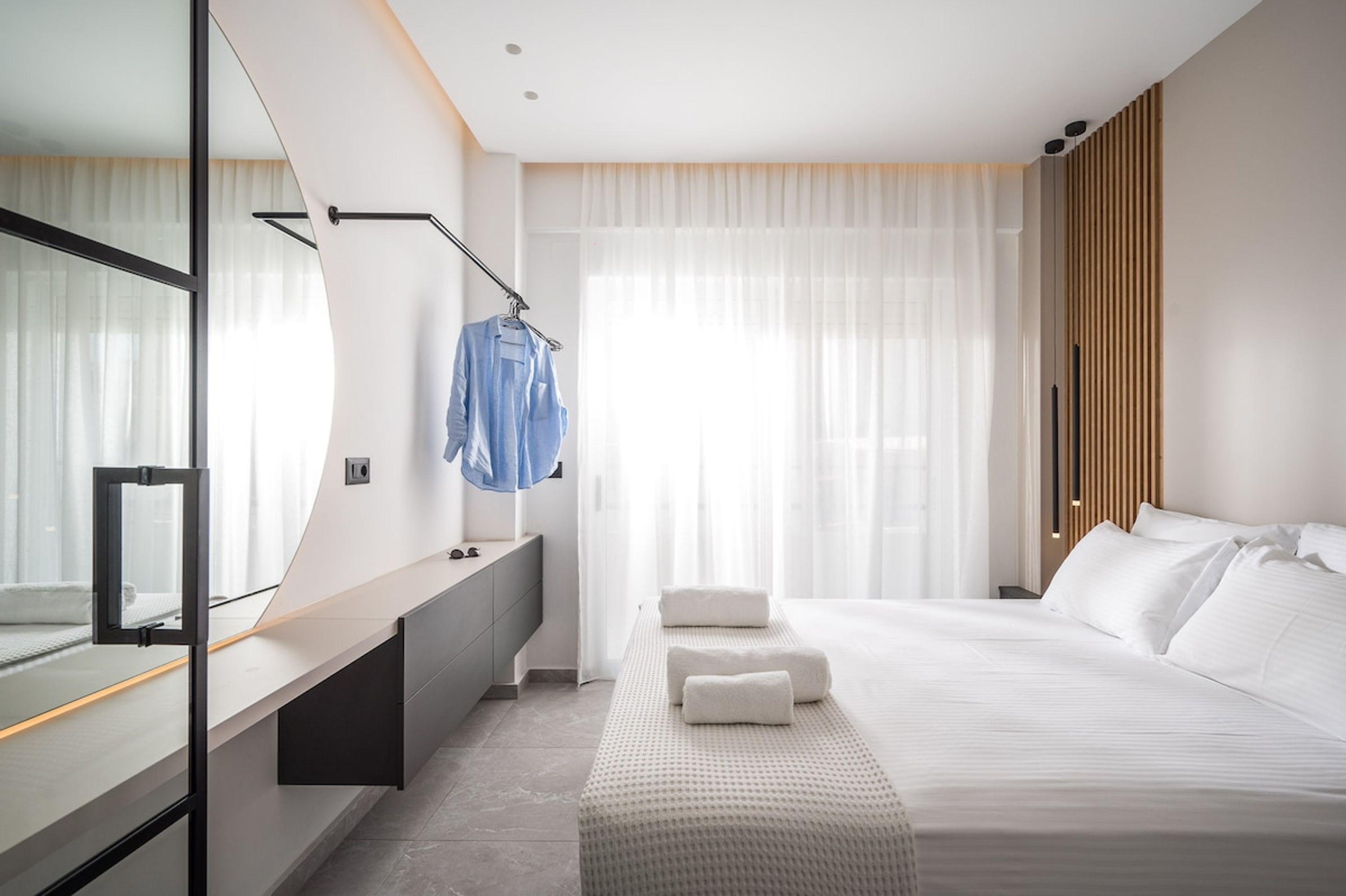 A modern hotel room with a large bed, a bedside table, a mirror, and a window with sheer curtains.