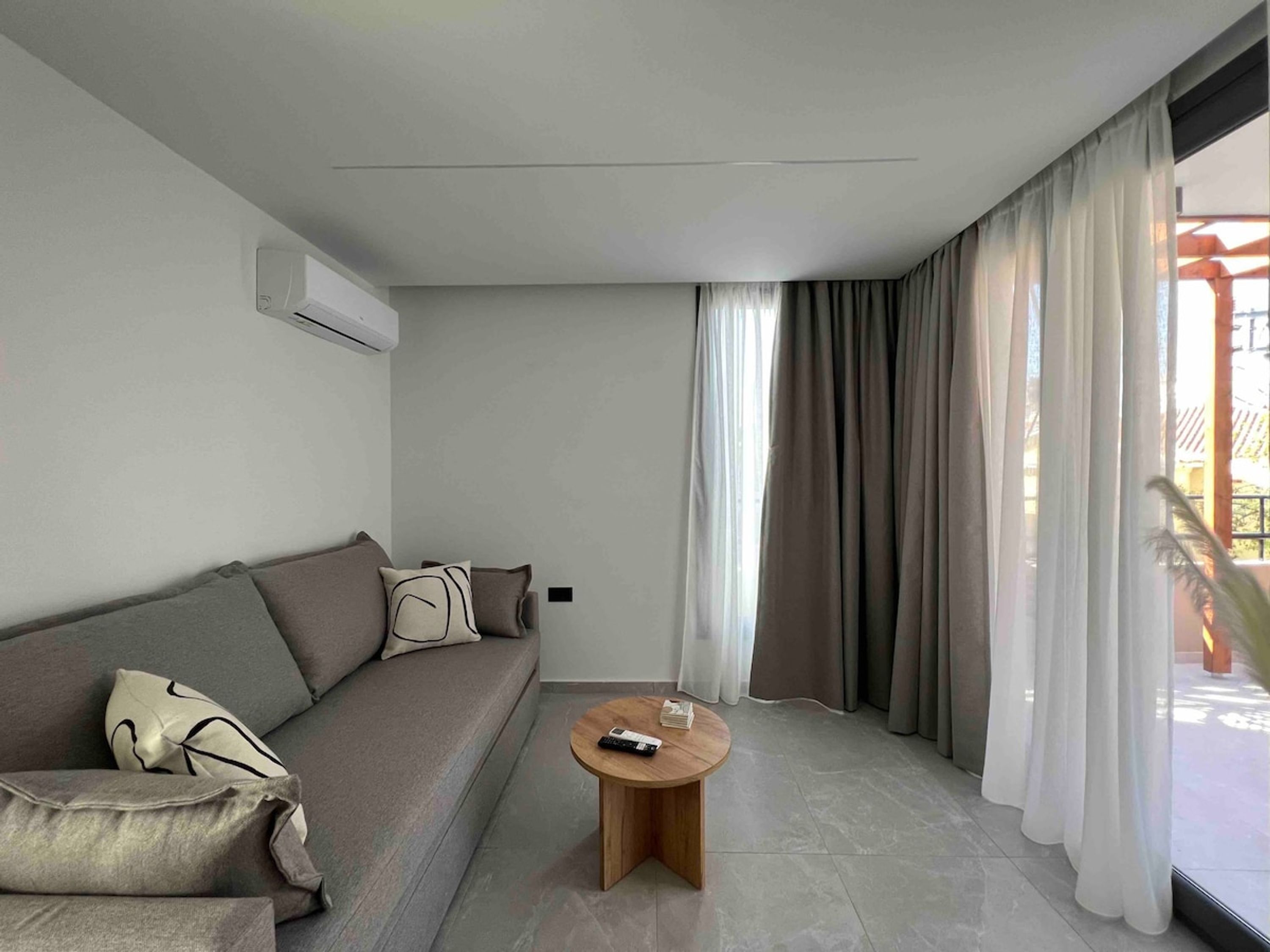A modern living room with a grey sofa, a round wooden coffee table, and a large window with curtains.