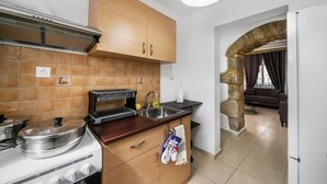 A kitchen with wooden cabinets, a stove, and a sink. There is a stone archway leading to a living area with a sofa and a table. - Mela s Home Agioi Deka (Gortyna)