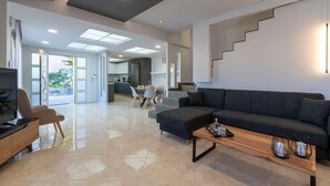 A spacious living area with a dark sofa, a wooden coffee table, and a TV on a wooden stand. A staircase leads to an upper level. - Villa Marie Nicol Private Pool (Malevizi)