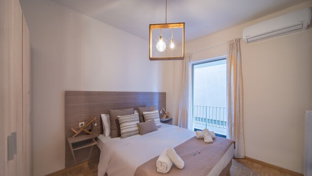A hotel room with a large bed, wooden headboard, bedside tables, a hanging light fixture, a window with curtains, and an air conditioning unit. - City Center Apartment Project by Cretan Houses (Heraklion)