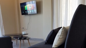 A modern living room with a flat-screen TV mounted on the wall, a dark sofa with cushions, a small round table with a tray of breakfast items, and a window with sheer curtains. - City Center Apartment Project by Cretan Houses (Heraklion)